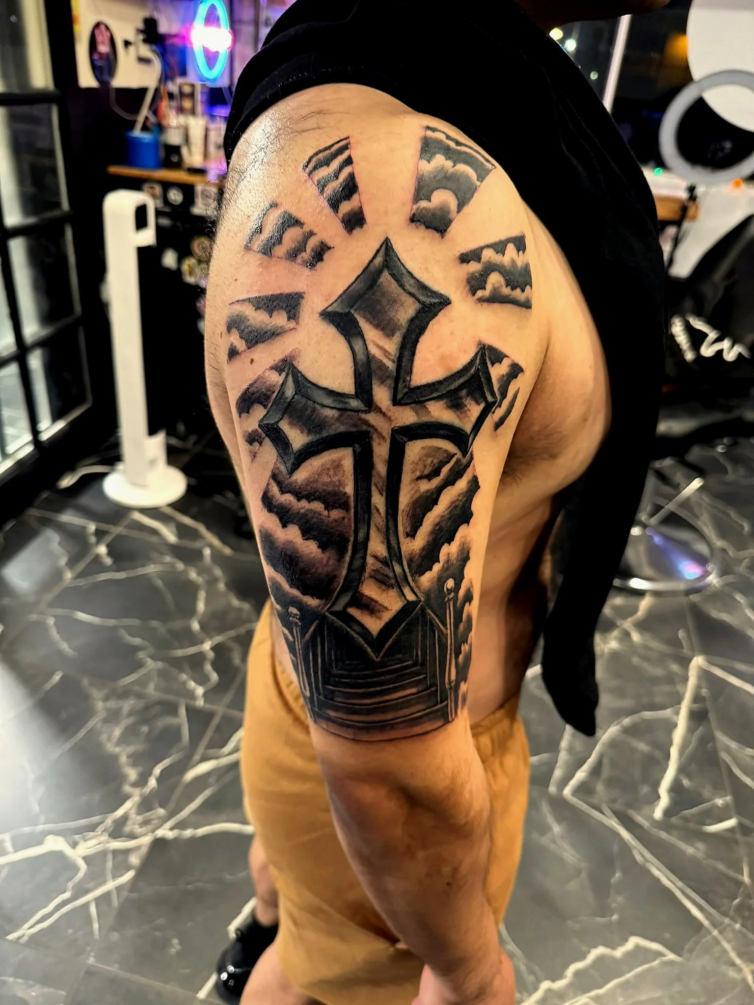 Tattoo on upper arm featuring bold black Gothic-style lettering, surrounded by dark, cloudy sky design, with a diamond shape at the center.
