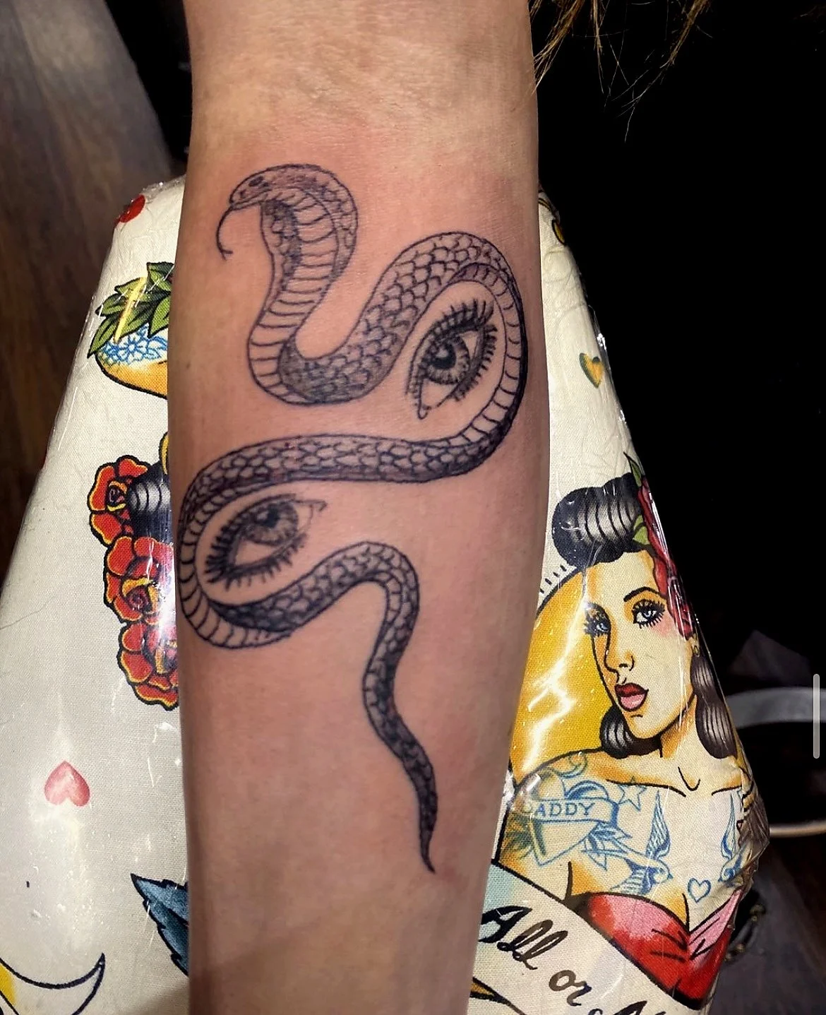 Tattoo of a snake with eyes on its coils on a person's forearm, with part of a cartoon-style tattooed woman with dark hair and a tattoo of a heart and banner visible on skin under the arm.