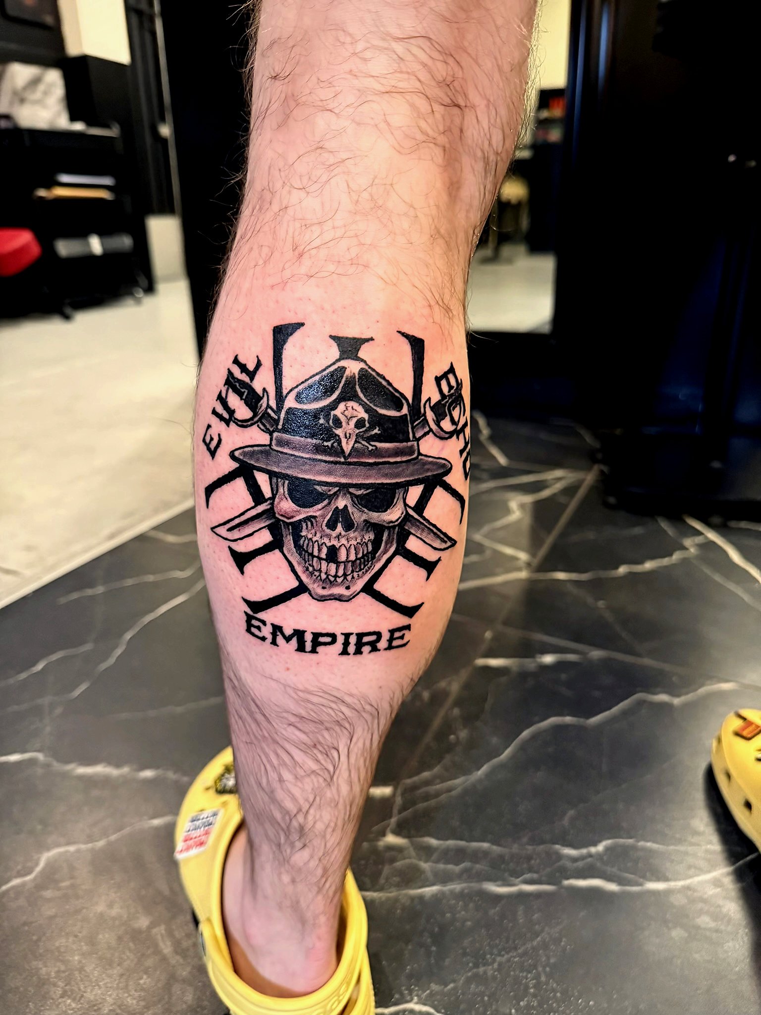 Tattoo on person's lower leg featuring a skull with a wide-brimmed hat, crossed katana swords behind it, and the words 'HUE' and 'EMPIRE' around the design.