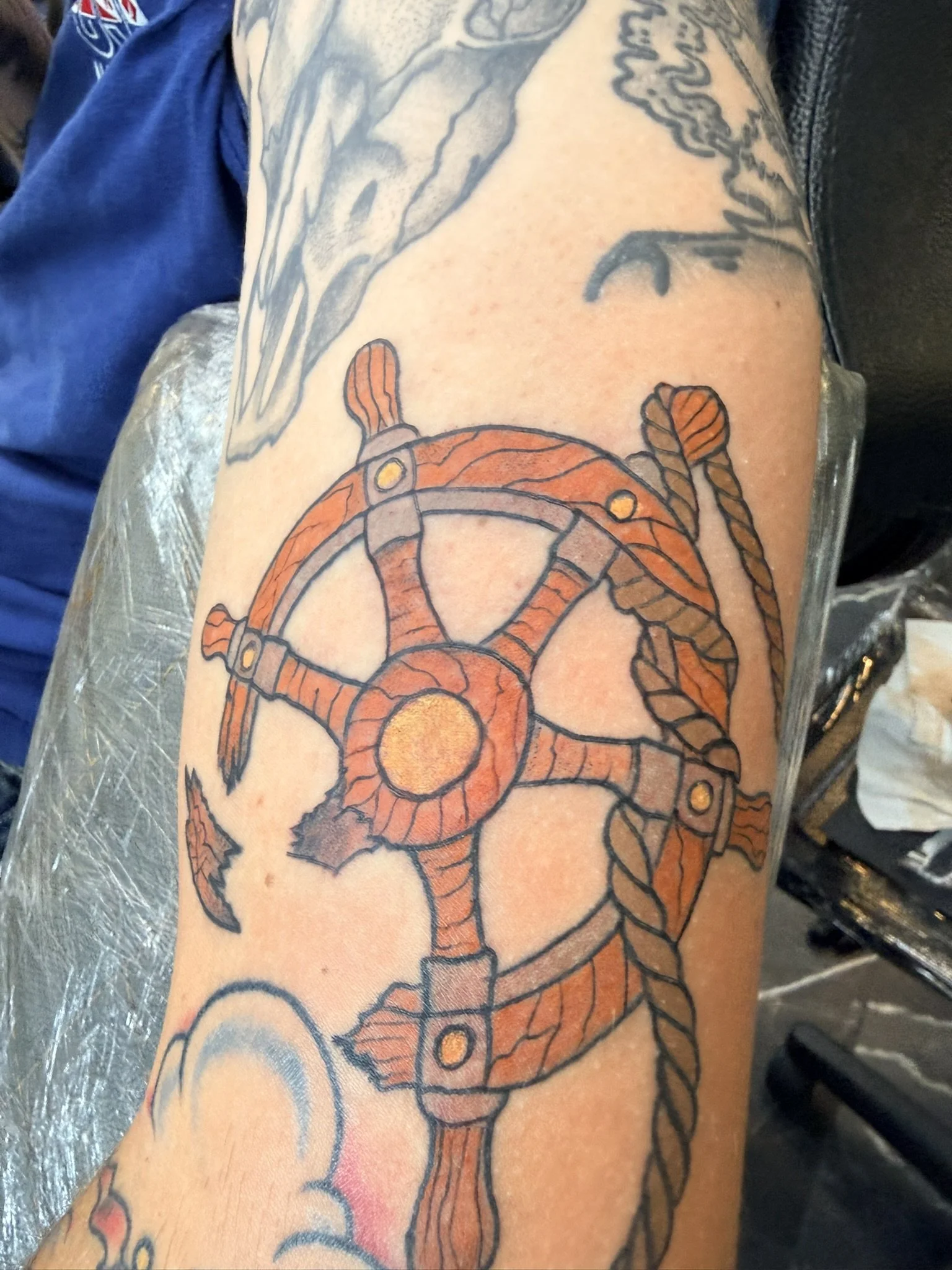 Close-up of a person's arm featuring a tattoo of a traditional-style orange and brown compass intertwined with a rope, on the upper arm.