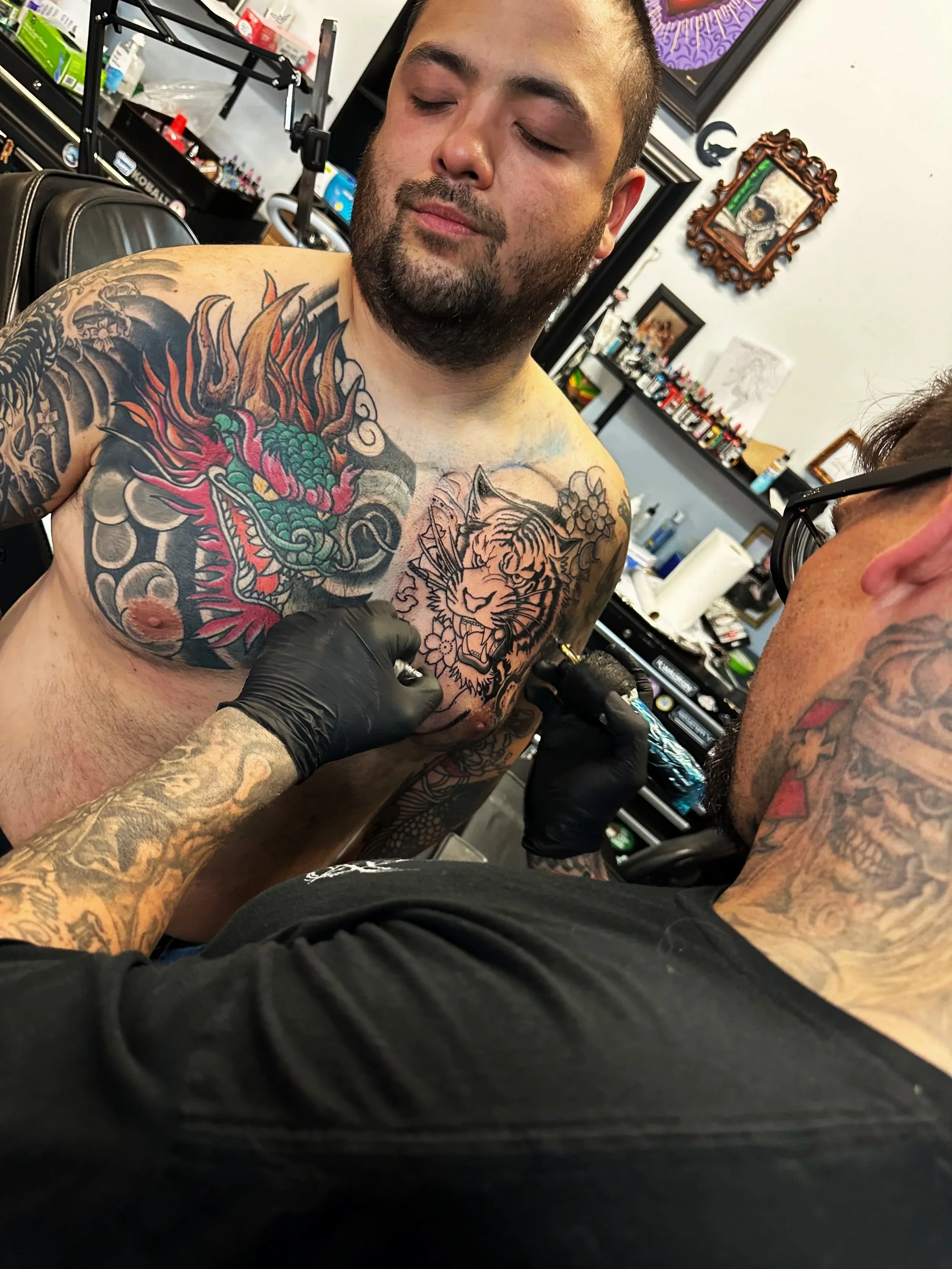A man with tattoos on his chest and arms sitting in a tattoo shop, getting a tattoo of a tiger's face on his chest from a tattoo artist wearing black gloves. The man has closed eyes and a slight smile, and another person with glasses is partially visible on the right side of the image.