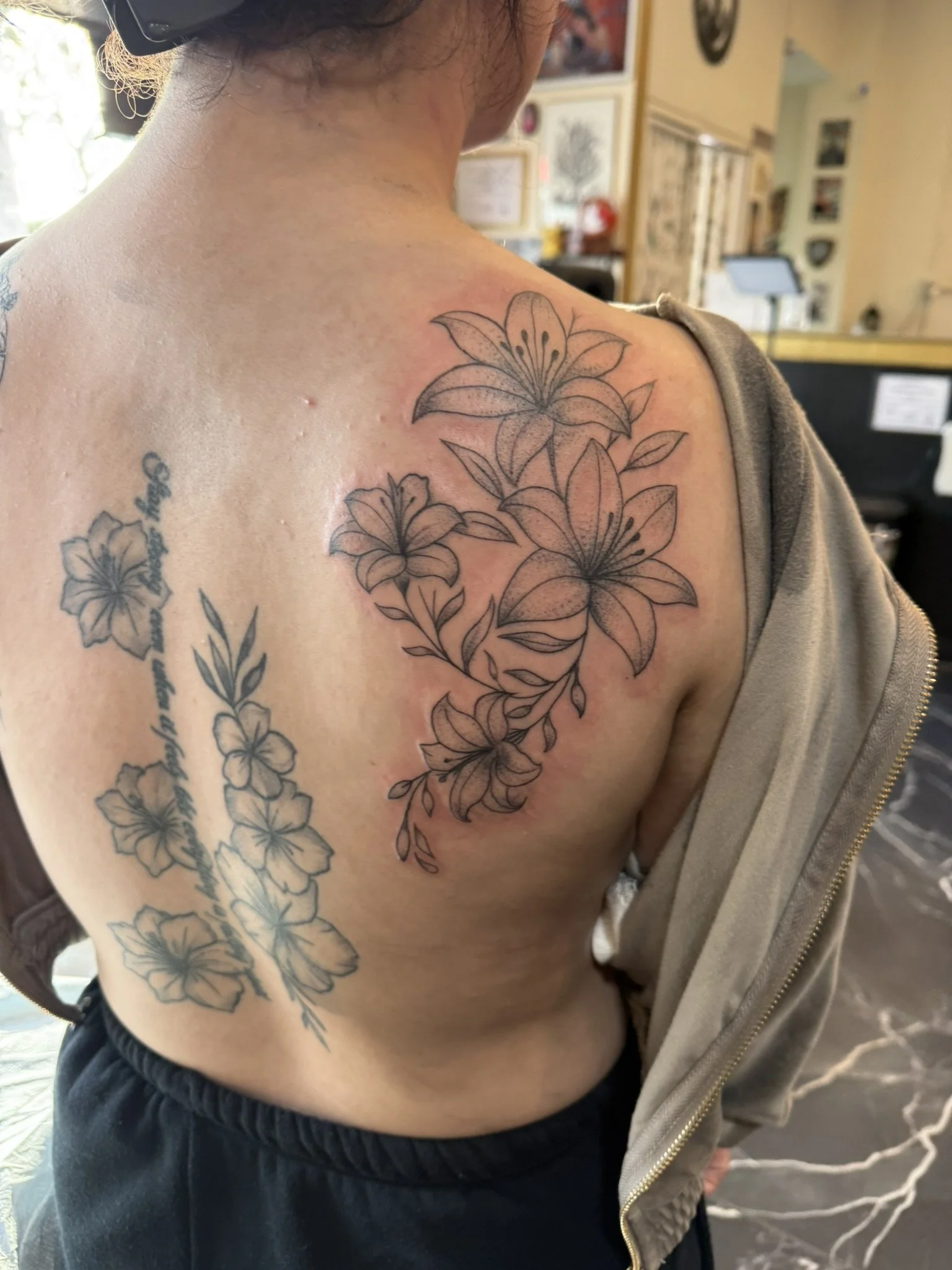 A person's upper arm and shoulder with multiple floral tattoos, including lilies and other flowers, in black ink. The person is wearing a gray hoodie with the zipper partially open, and background includes framed artwork on a wall and some furniture.