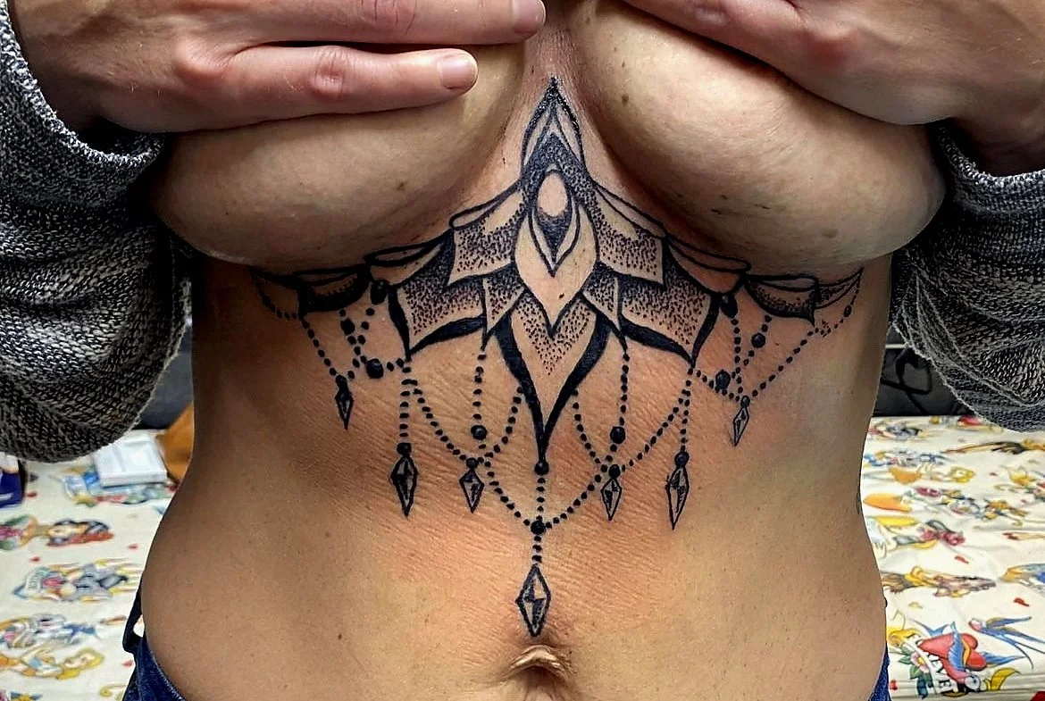 Close-up of a person's chest with a large, detailed black tattoo design that extends from the upper chest to the mid-abdomen, featuring a symmetrical, ornamental pattern with hanging bead and diamond-shaped elements, and hands covering the breasts.