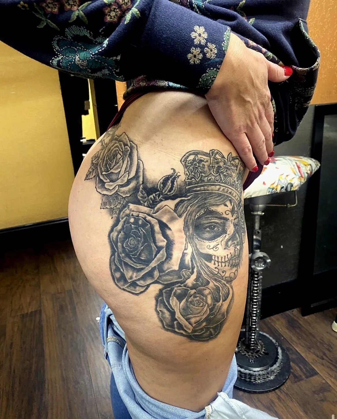 Tattoo of a skull face with a crown, surrounded by roses, on a woman's thigh.