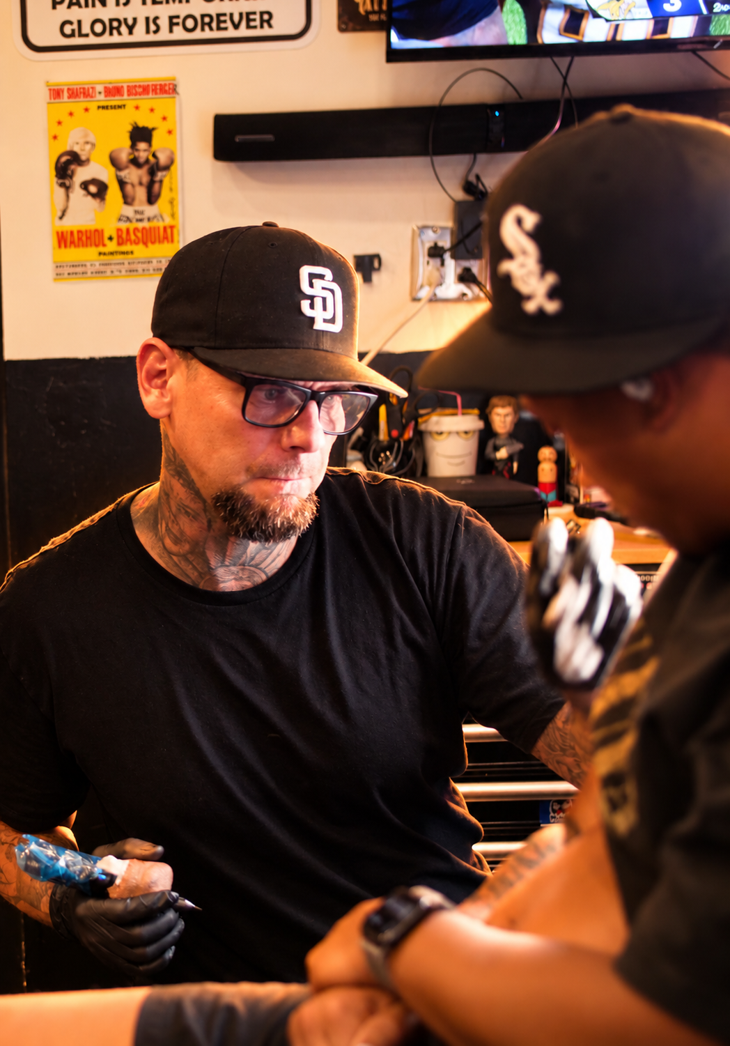 A tattooed man wearing sunglasses and a black tank top standing outside a storefront with orange signage that reads 'TATTOO INK SPOT'.