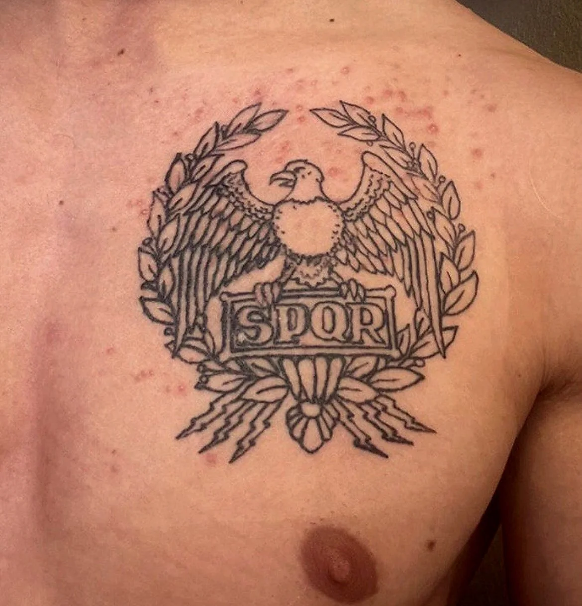 Tattoo of an eagle with outstretched wings, holding a shield with the letters "SPQR" on it, surrounded by a laurel wreath, on a person's chest.