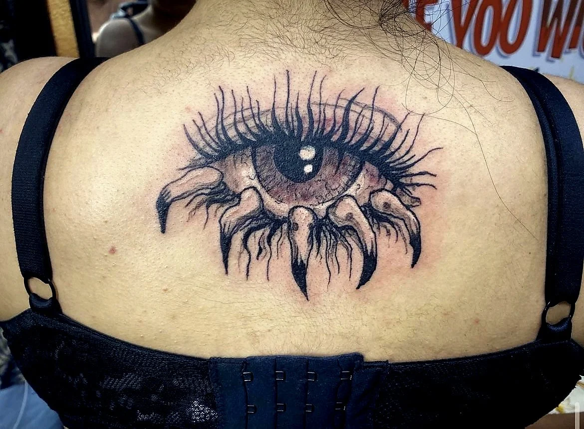 Tattoo of a realistic human eye with long eyelashes, with spider legs extending from the lower eyelid, on a person's upper back.