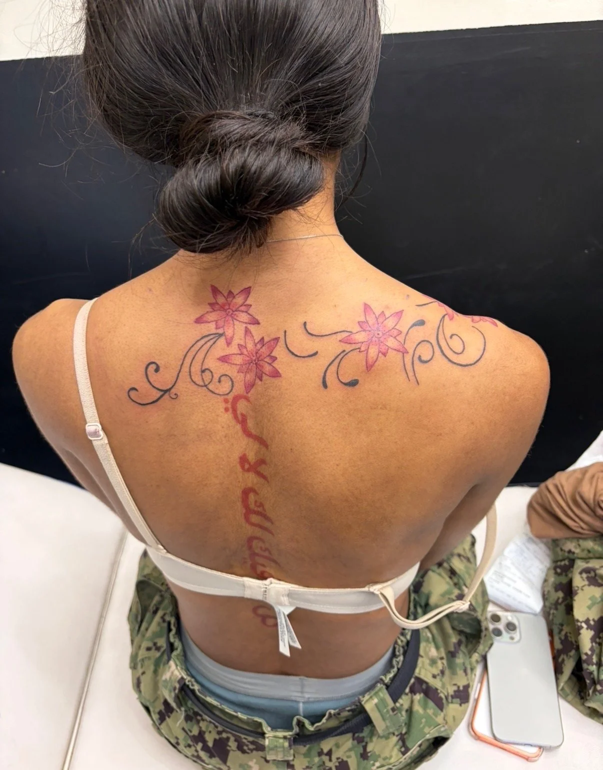 Back of a woman with dark hair styled in a bun, displaying a tattoo of pink flowers and black swirls on her upper back and a red script running vertically down her spine. She is wearing a white bra and camouflage pants, seated on a white surface with