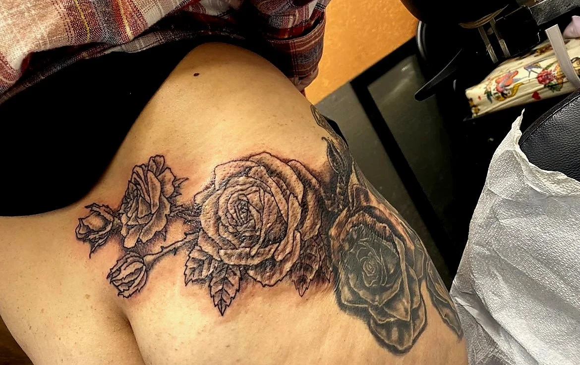 A person's shoulder with a freshly inked black and gray tattoo of roses and leaves.