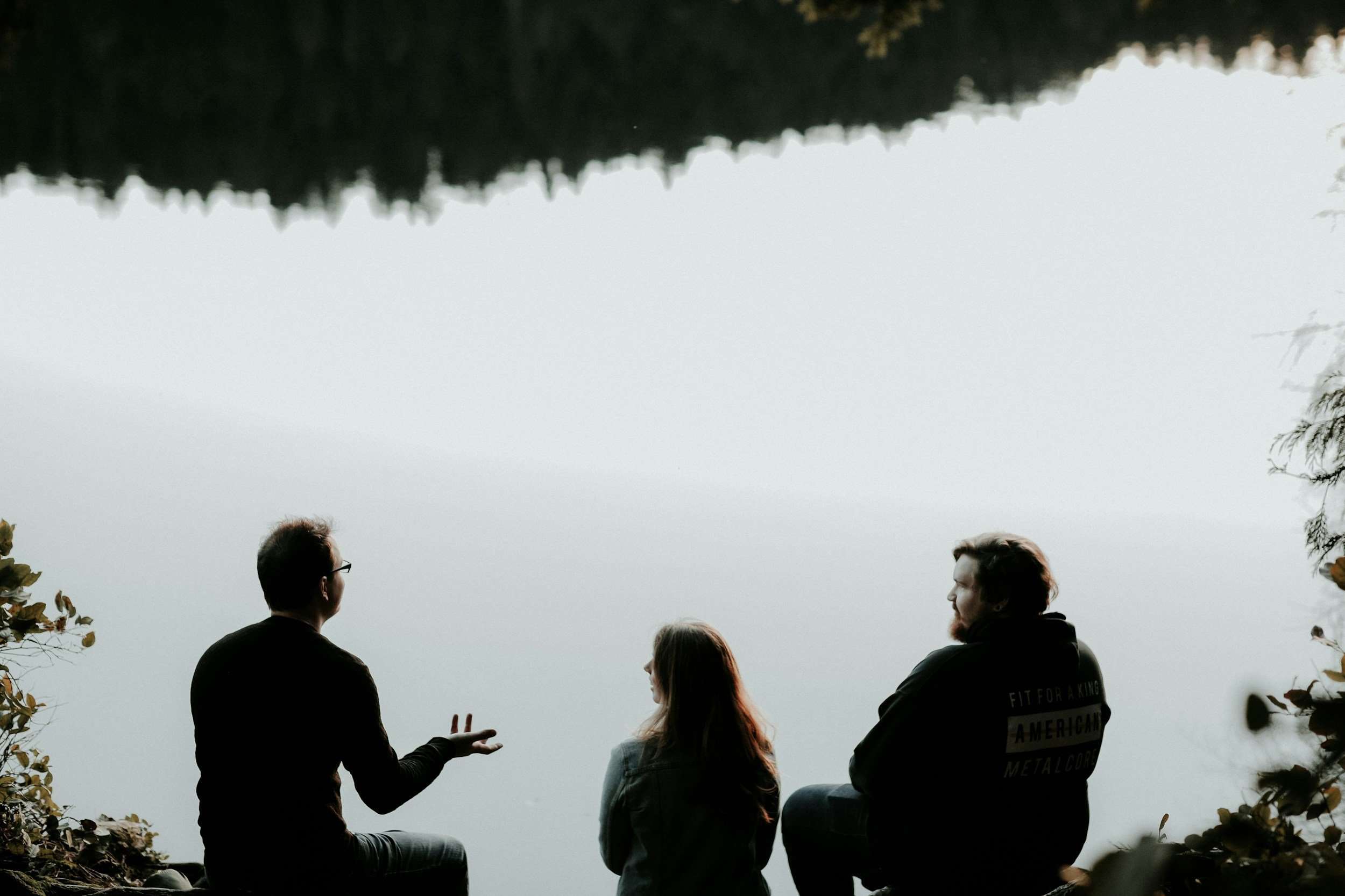 People sitting outdoors near water with trees in the background, two men are visible in the foreground wearing dark jackets, a woman with long hair is talking to one of them, and another person is partially visible on the right. The sky appears overcast.