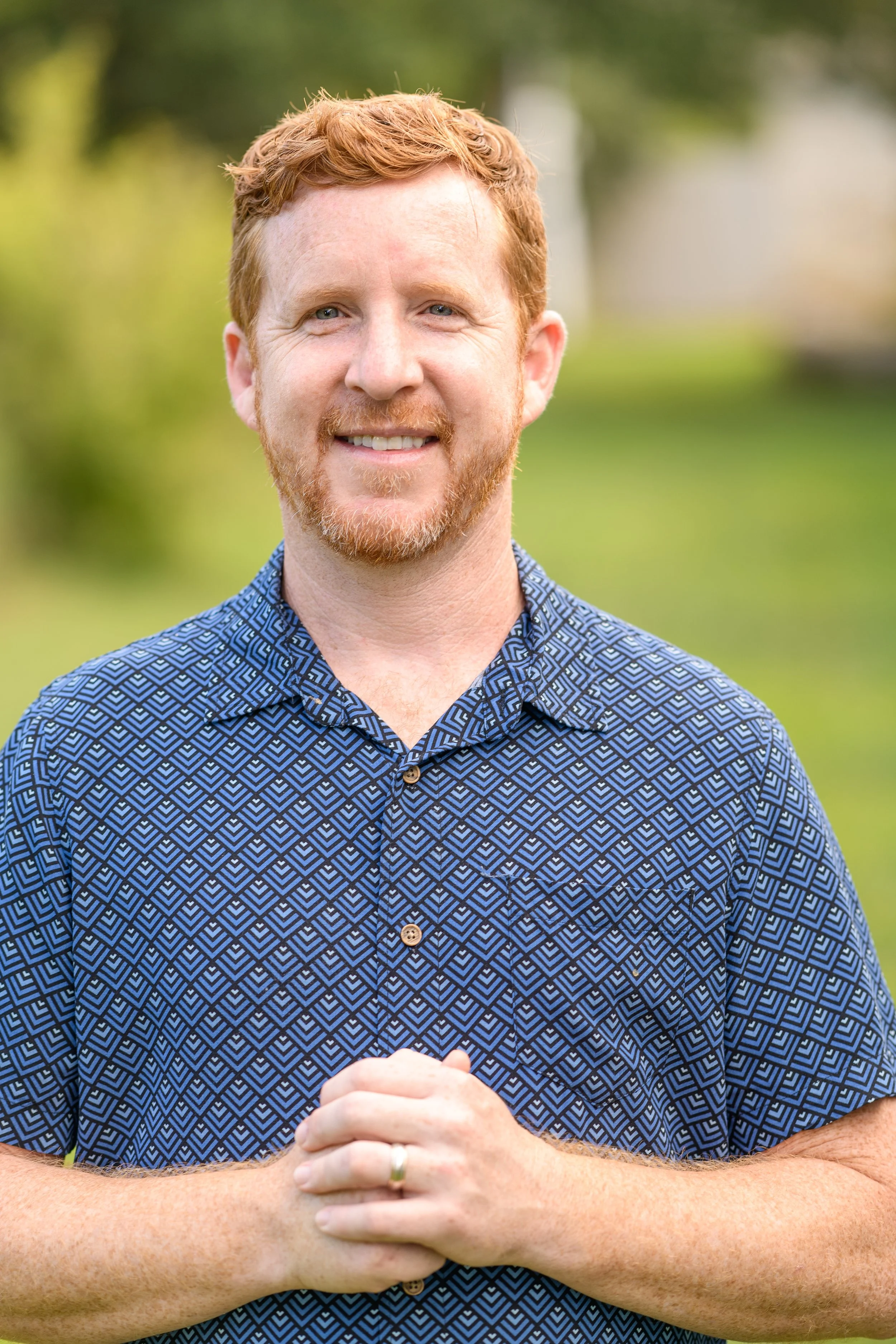 A man with red hair and a beard smiling outdoors, wearing a patterned button-up shirt, standing with hands clasped.