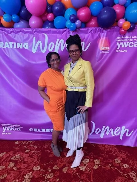Two women standing in front of a purple backdrop decorated with balloons, at a Women's celebration event. One woman is wearing an orange dress, and the other is dressed in a yellow blazer with a black and white pleated skirt.
