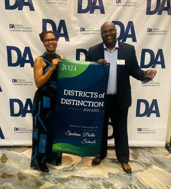 A woman and a man are standing together at an awards event, holding a large green and blue banner that reads "2024 Districts of Distinction Award proudly presented to Spokane Public Schools." They are smiling and dressed in formal attire, with a backdrop featuring the District Administration Leadership Institute logo.
