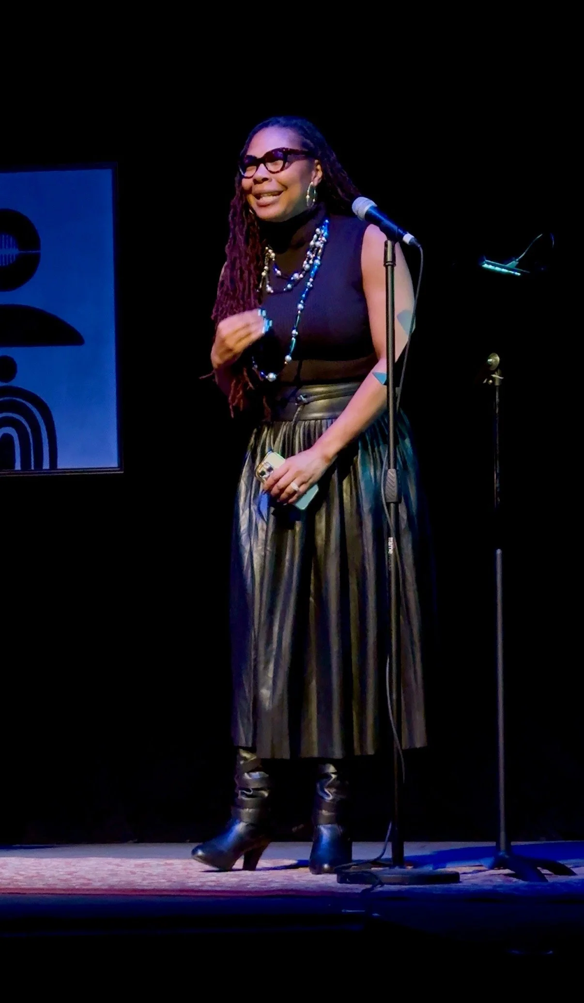 Woman with black hair, glasses, and multiple necklaces speaking into a microphone on stage, wearing a black sleeveless top, striped pants, and black boots.