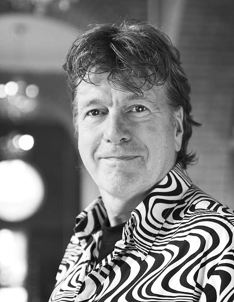 Black and white portrait of a man with wavy hair, smiling, wearing a patterned shirt with swirling designs.