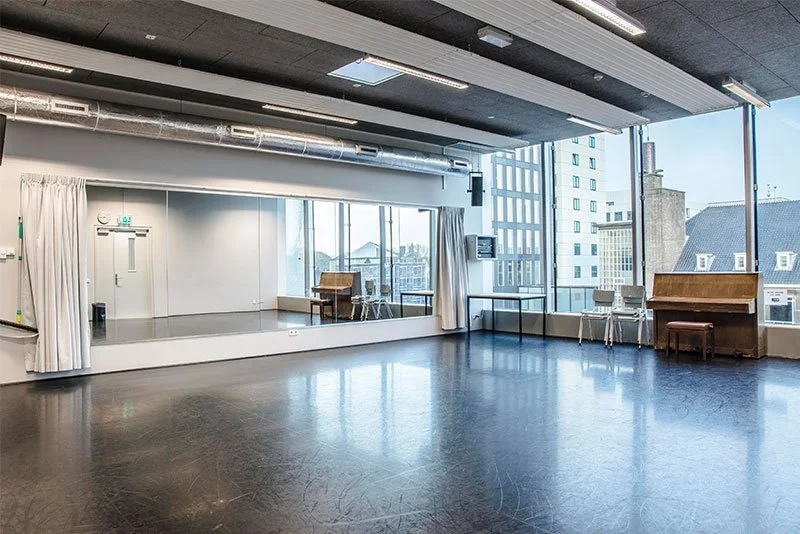 Empty dance or yoga studio with large windows, wooden piano, and minimal furniture.