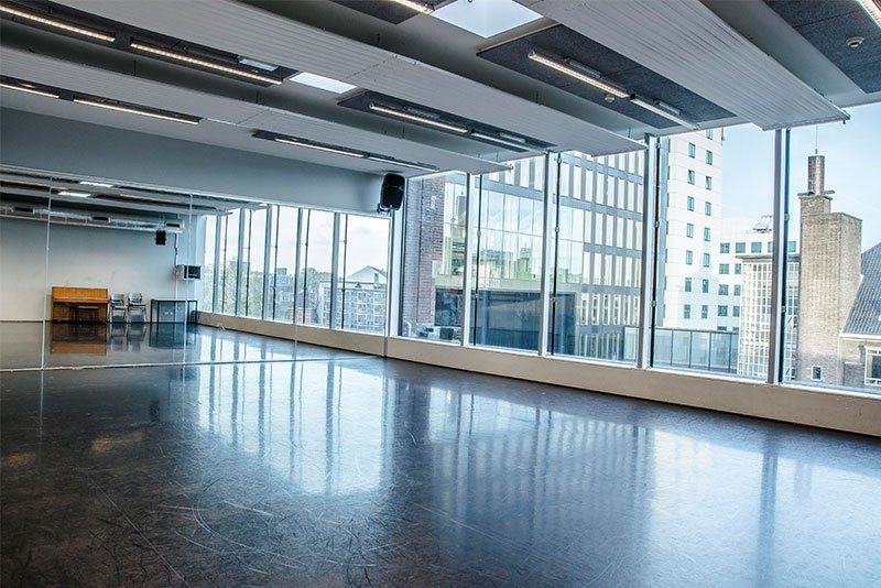 An empty, spacious dance or event studio with large floor-to-ceiling windows showing a cityscape outside, a few chairs and a table in the corner, and ceiling-mounted lights.