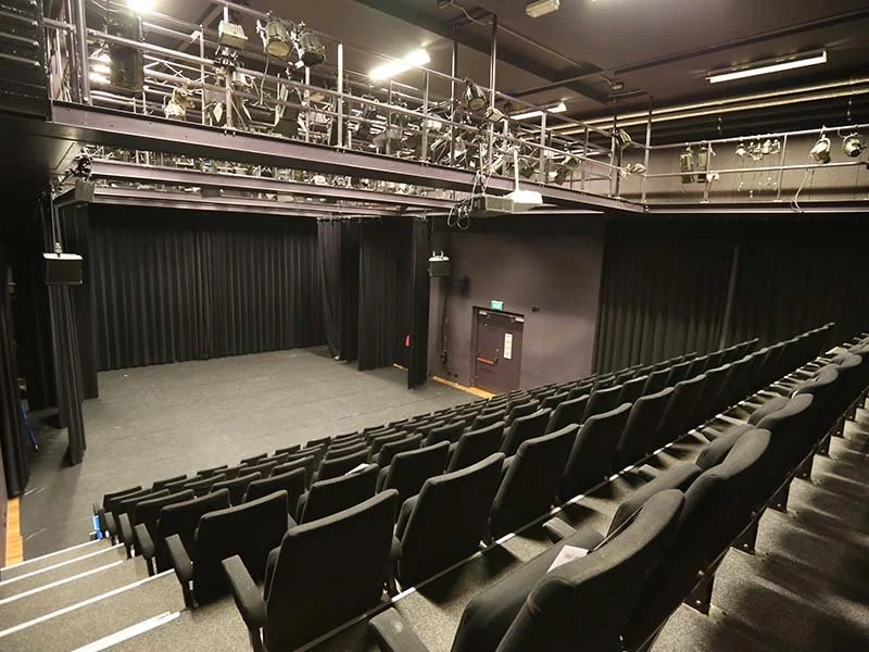 Empty black theater with a stage, black curtains, and theater seats arranged in ascending rows.