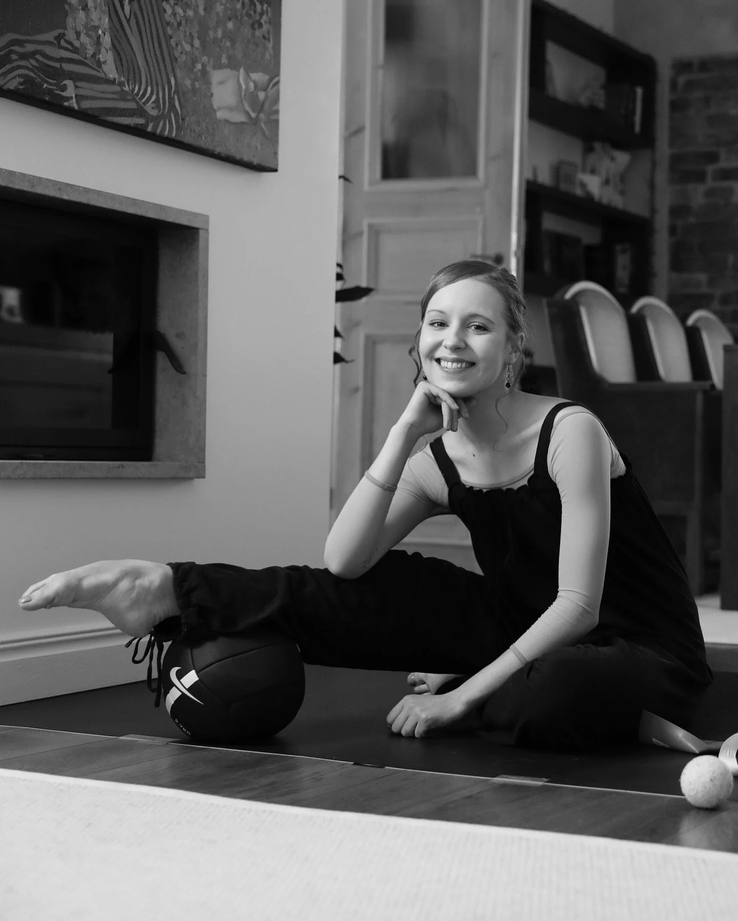 A young woman sitting on a yoga mat indoors, smiling, resting her chin on her hand, with a medicine ball under her leg and a small exercise ball nearby.