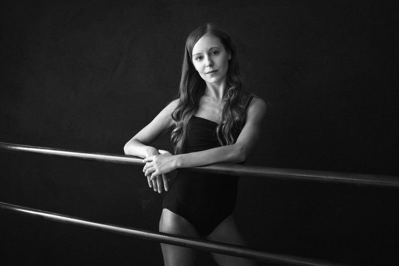 A woman in a black leotard standing behind a ballet barre against a dark background.