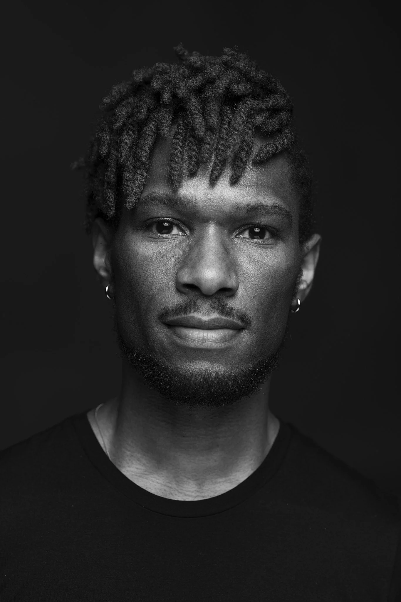 Black and white portrait of a young man with short dreadlocks, earrings, and a goatee, looking directly at the camera against a dark background.