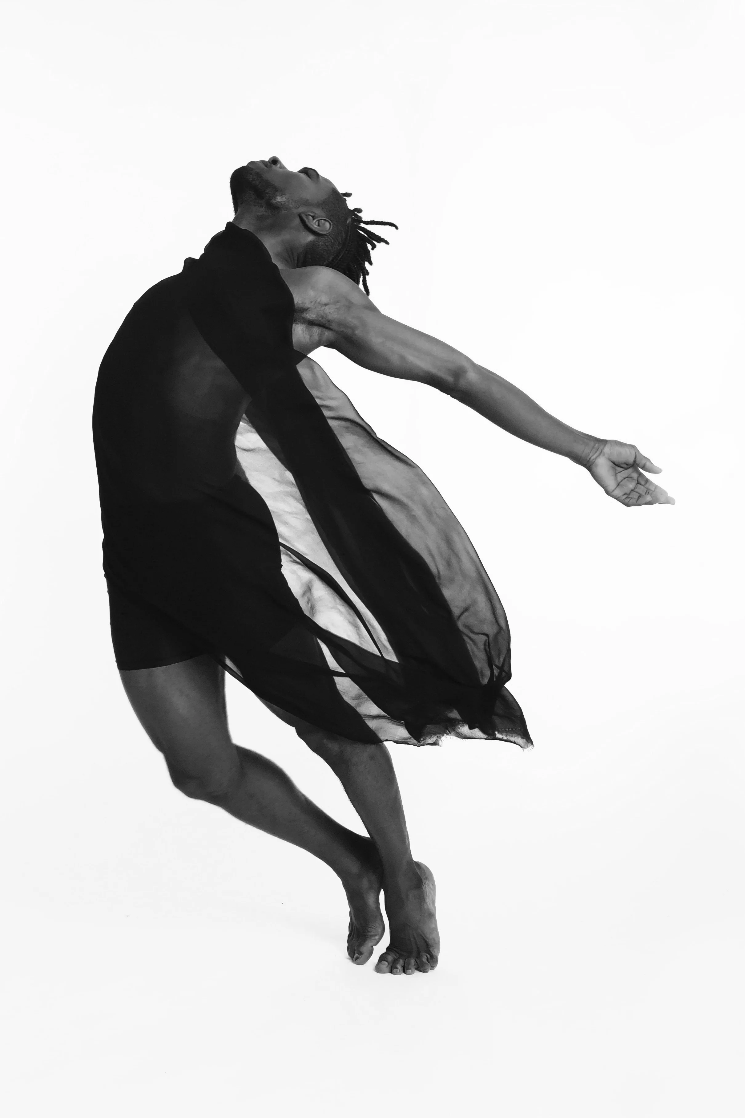 A person with dreadlocks wearing a flowing dress performing a dance move on a plain background.