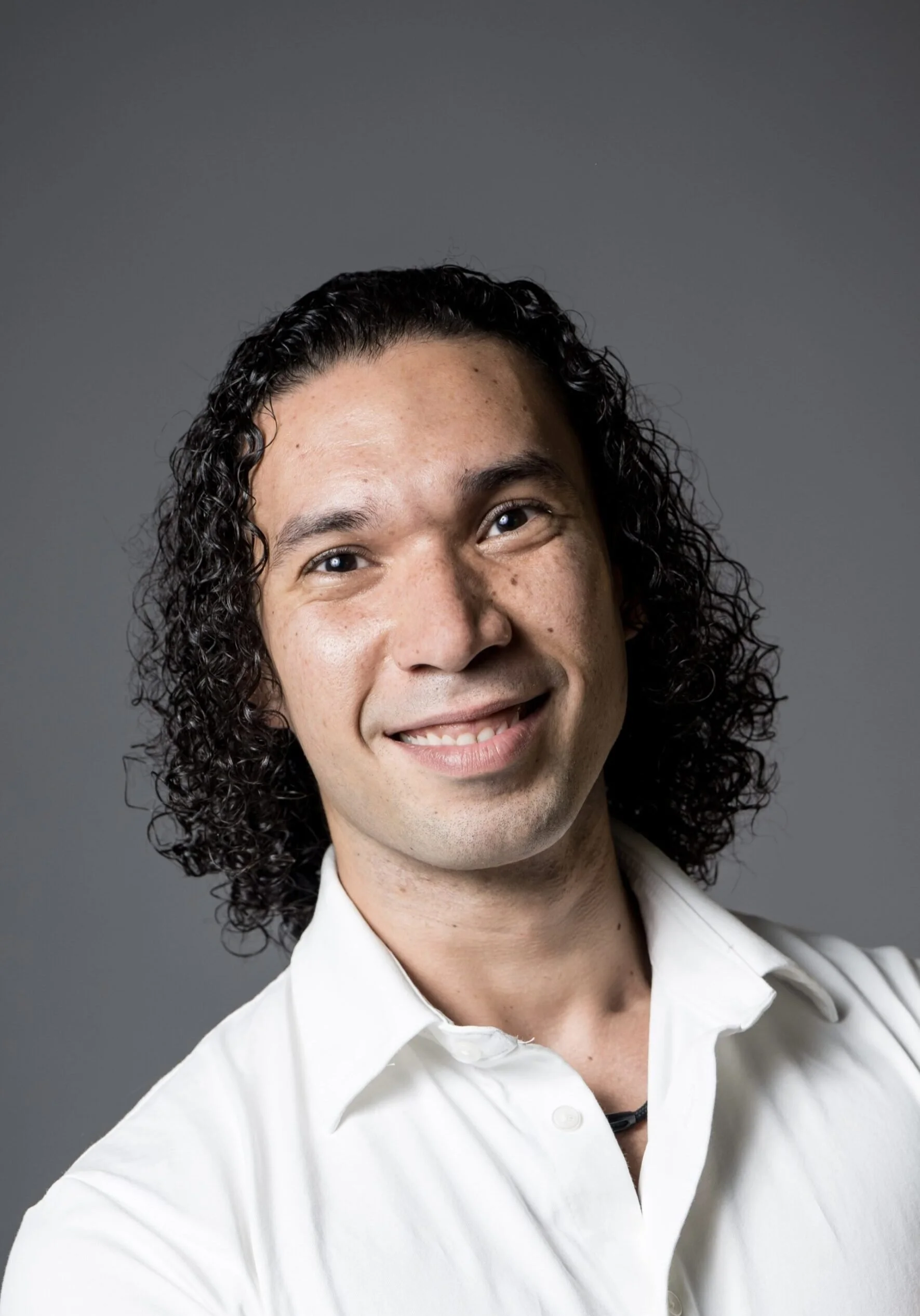 A young man with curly dark hair, wearing a white collared shirt, smiling and looking at the camera against a gray background.