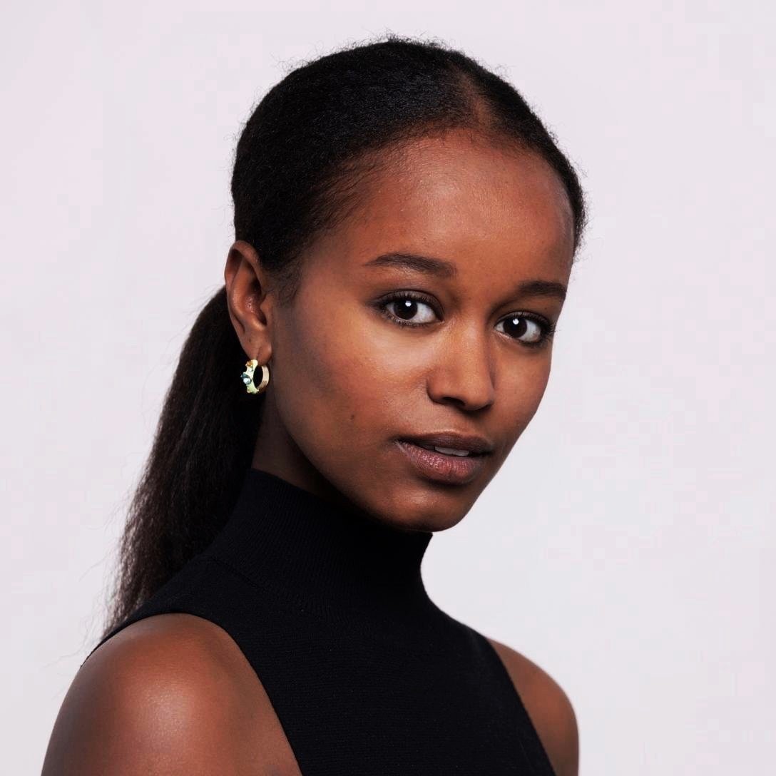A portrait of a young Black woman with dark hair pulled back, wearing a black sleeveless top and gold earrings, against a plain white background.