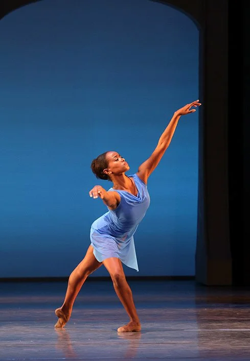 A female ballet dancer performing on stage in a blue costume, striking a pose with her right arm extended outward and left arm raised high, with a plain blue background.