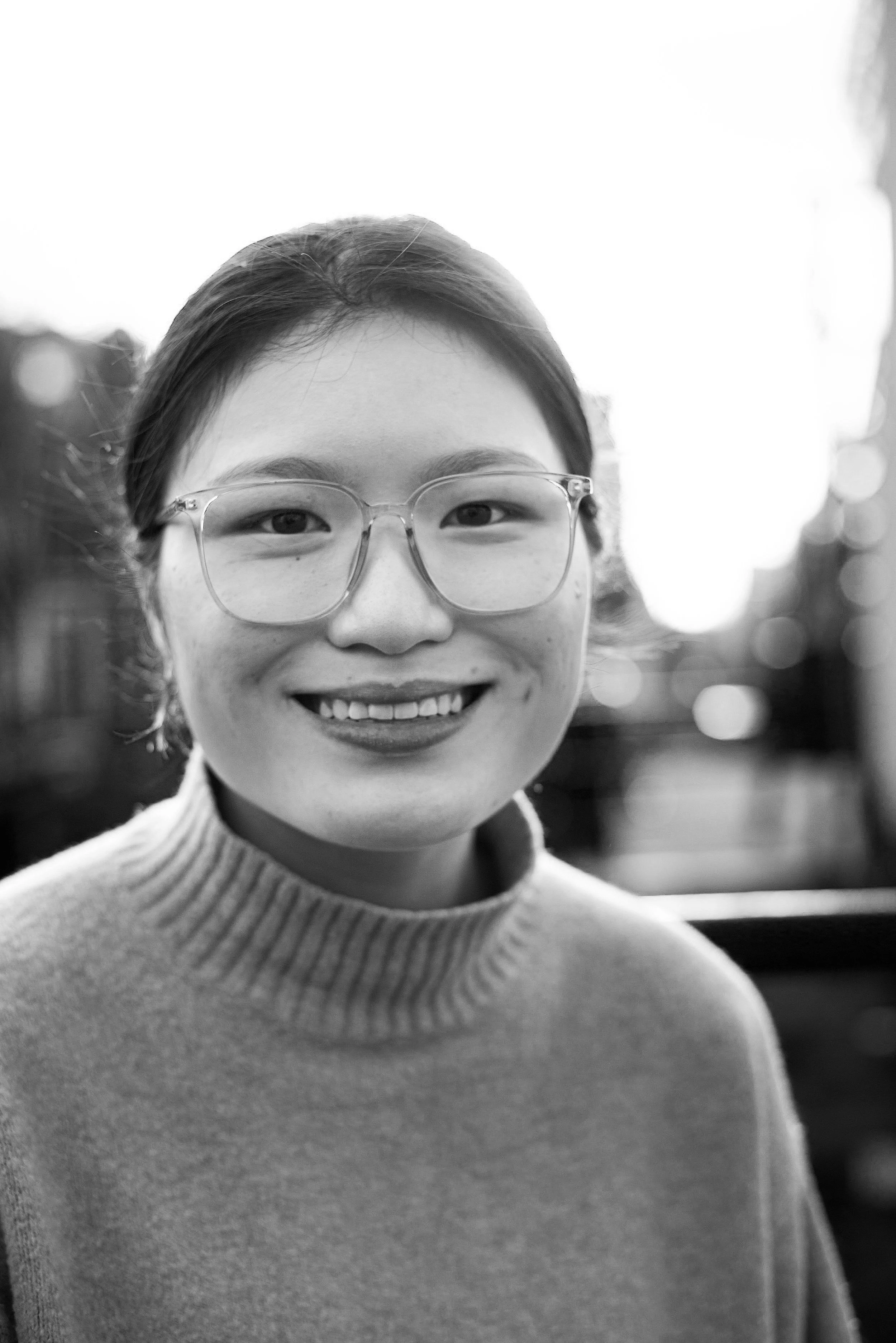 Black and white photo of a woman smiling outdoors, wearing glasses and a sweater.