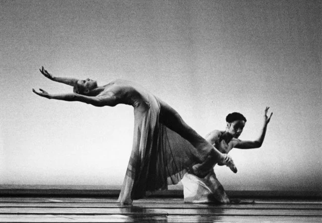 Two ballet dancers perform a pose on stage, with one dancer arching backward and the other supporting her, in a black and white photo.