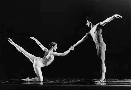 Two ballet dancers practicing on stage; the female dancer is on her knees with one arm extended backward and the other reaching forward, holding the male dancer's hand; the male dancer is standing with one arm extended outward and the other holding the female dancer's hand.