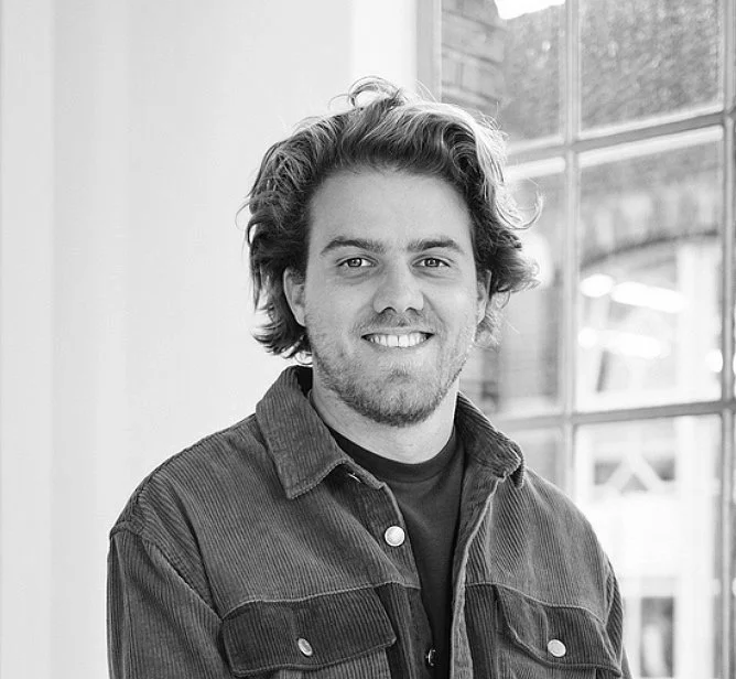 Black and white portrait of a young man with wavy hair, smiling, wearing a denim jacket, in front of large windows.