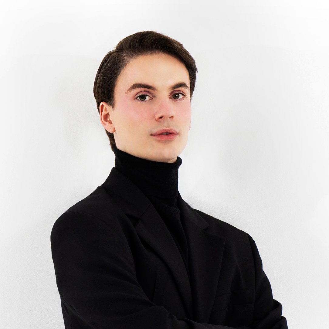 Portrait of a young man with dark hair wearing a black turtleneck and jacket, standing against a plain white background.