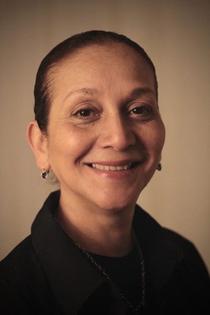 A woman with slicked-back hair, smiling, wearing earrings and a black top, against a plain background.