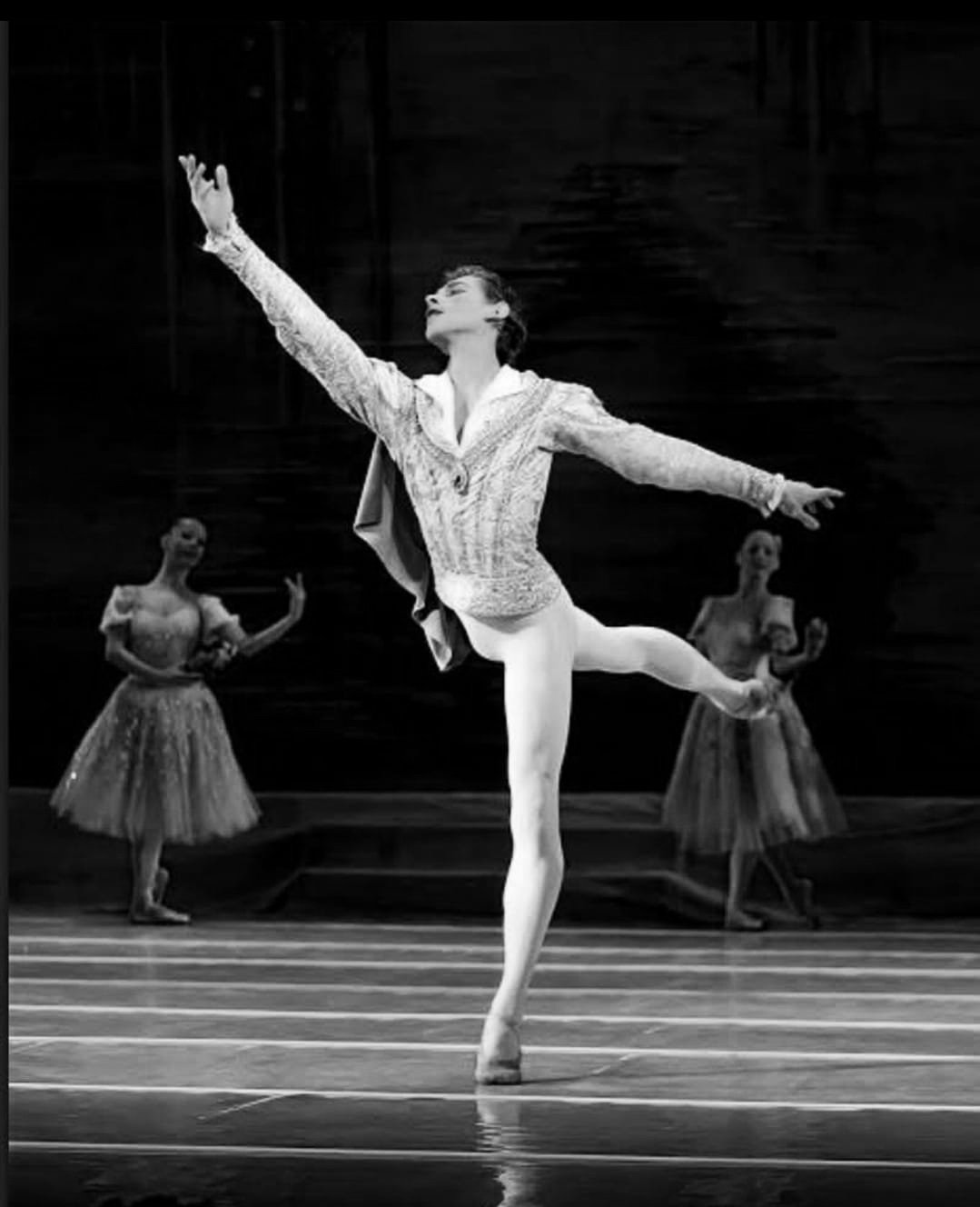 A ballet dancer performing a pose on stage with two dancers in tutus in the background, in black and white.