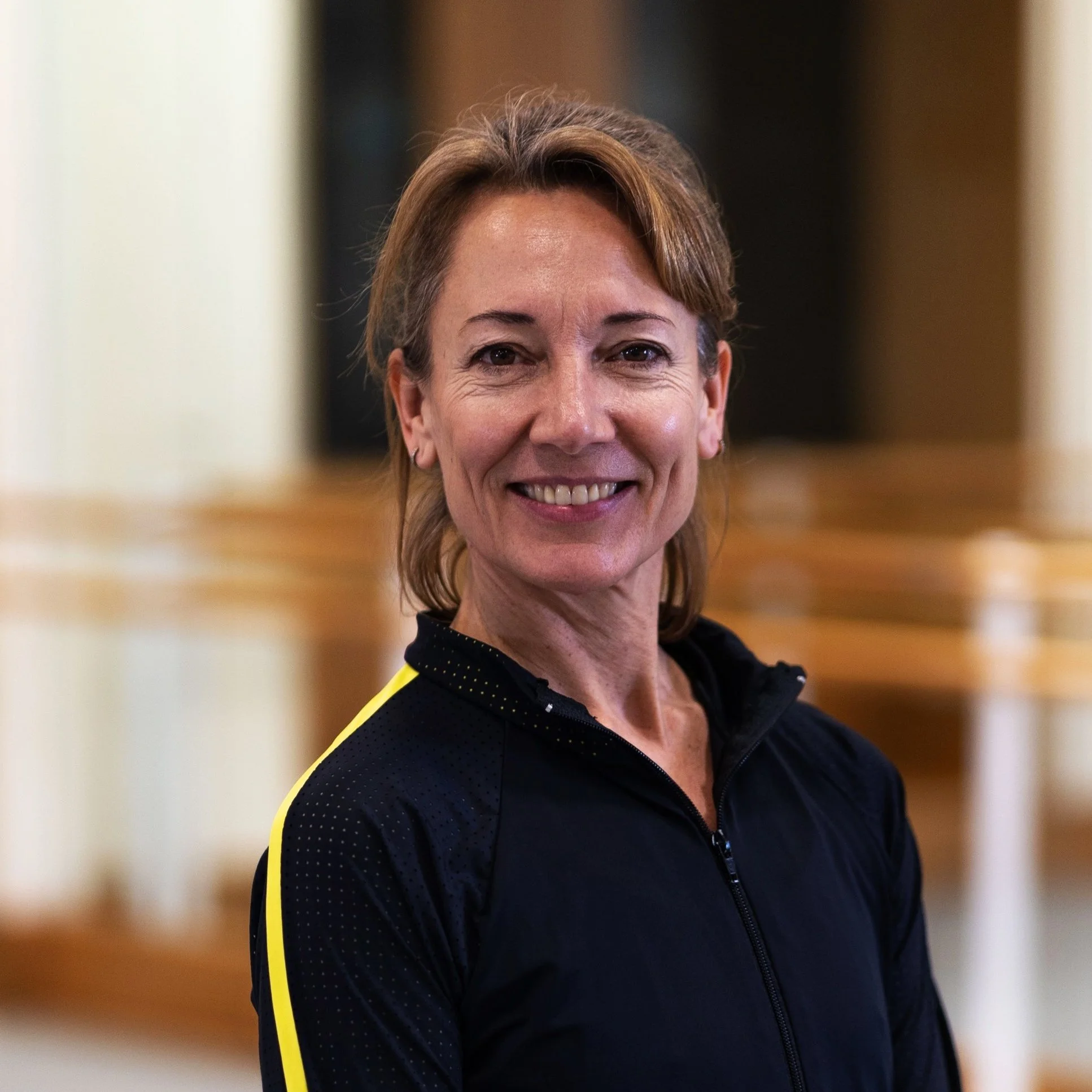 A middle-aged woman with shoulder-length light brown hair smiling, wearing a black athletic jacket with yellow accents, indoors with wooden architectural elements in the background.