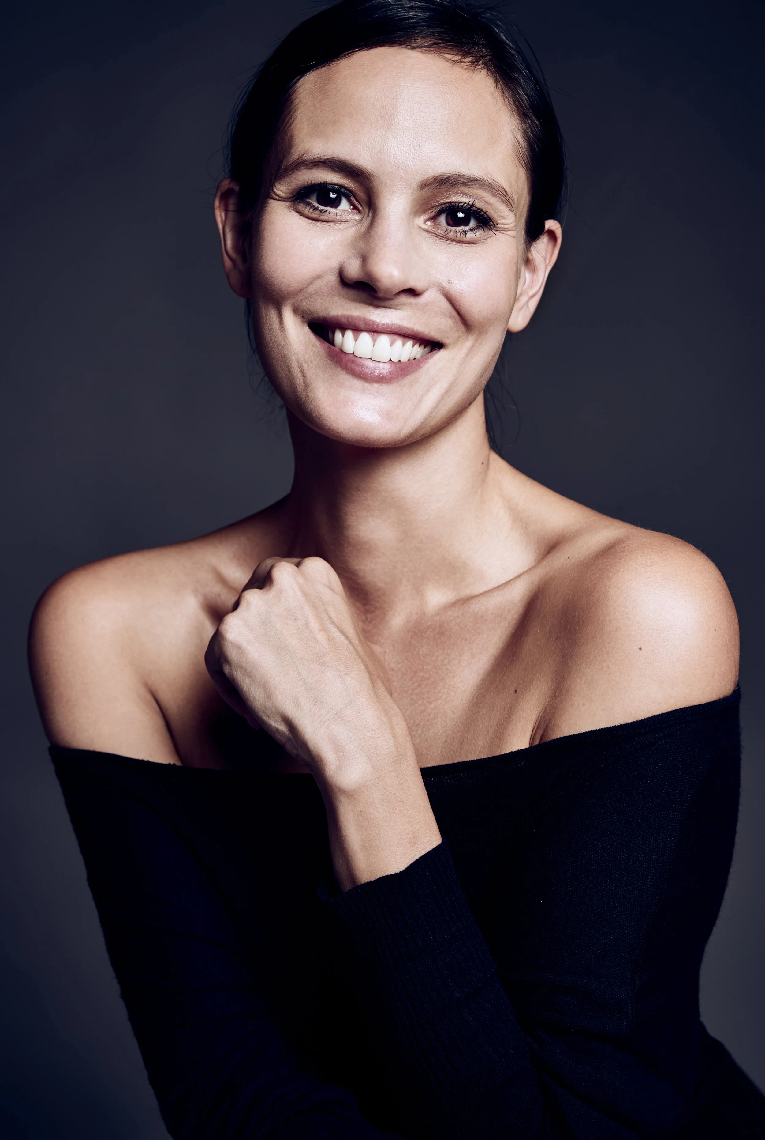A smiling woman with dark hair, wearing a black off-shoulder top, posing against a dark background.