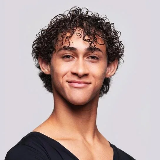 A young man with curly dark hair smiling and wearing a black shirt, against a light gray background.