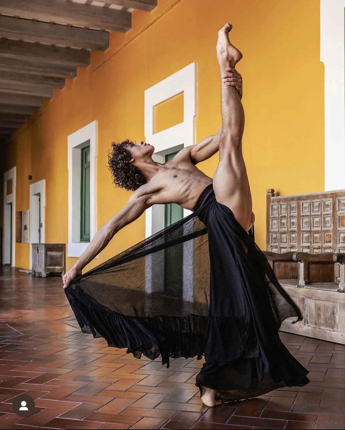 A man with curly hair, shirtless, wearing a black flowing skirt, performing a dance pose in a room with yellow walls, white window frames, and wooden flooring.