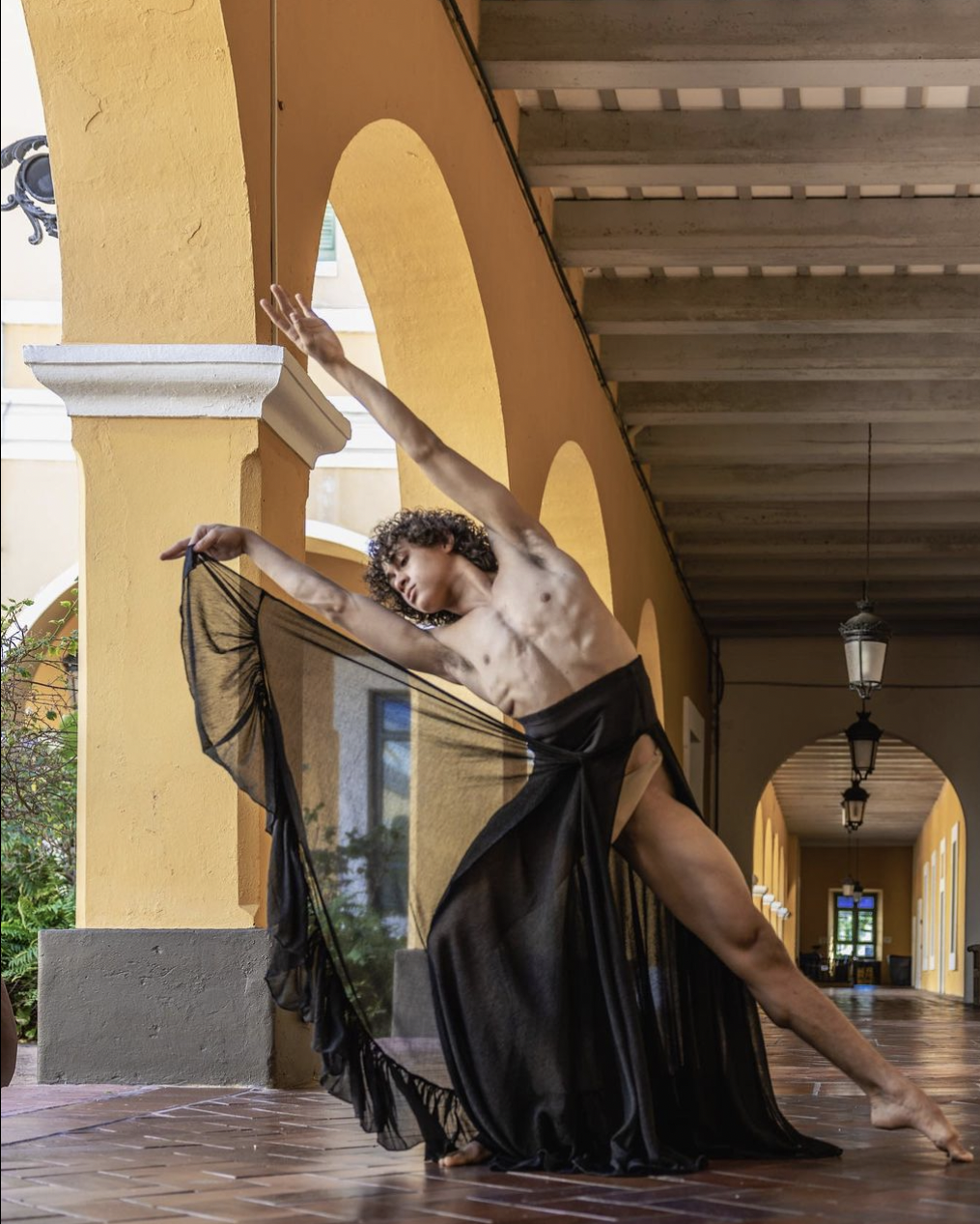 A shirtless person with curly hair, dancing in a corridor with yellow walls and brick flooring, wearing a black sheer skirt, posing with arms extended and one knee bent.