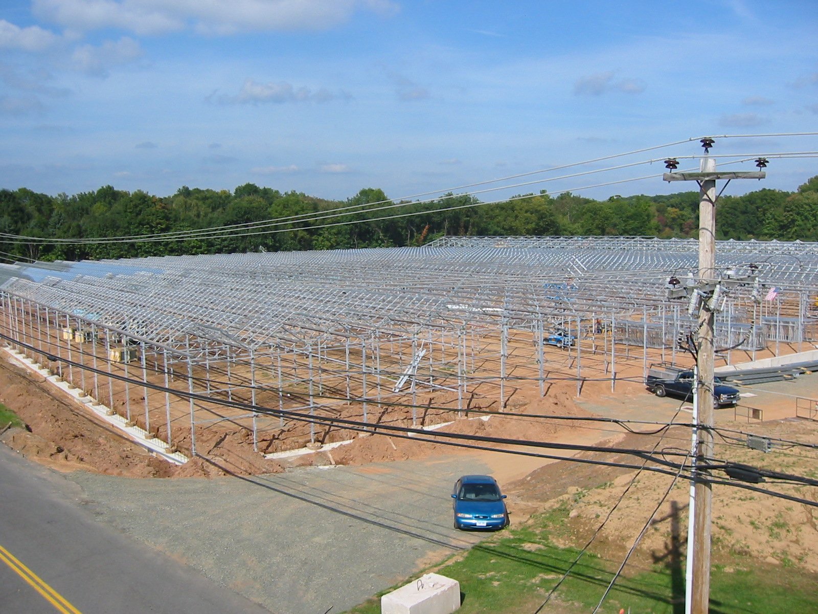 Large solar panel installation with metal framework and support structures, power lines and utility pole in the foreground, and a background of trees and blue sky.