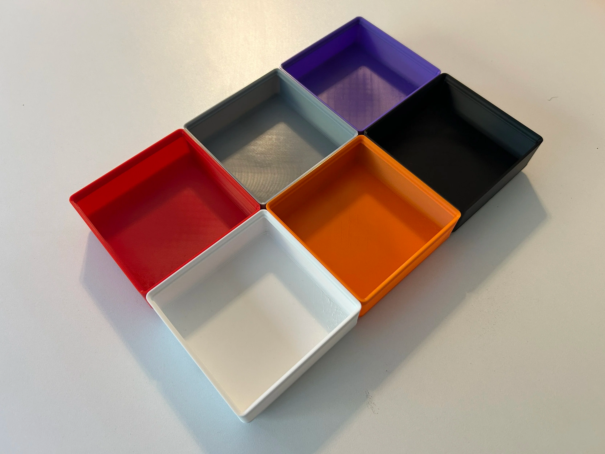 A top view of six empty rectangular plastic containers in red, gray, purple, black, orange, and white, arranged on a white surface.