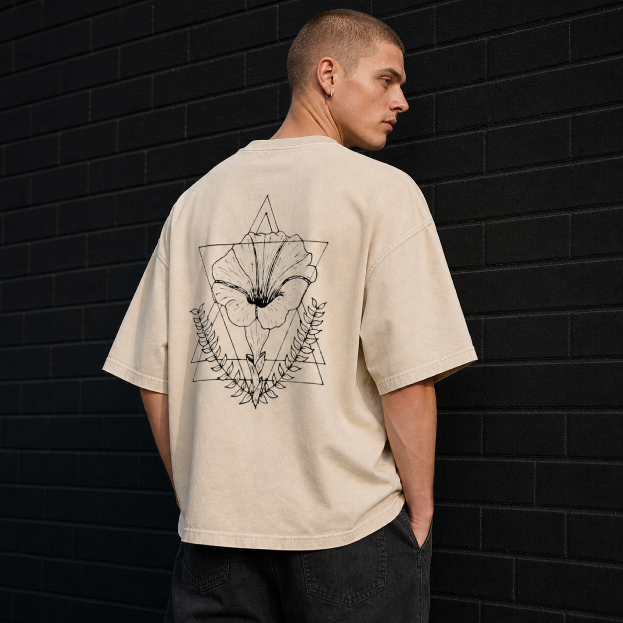 A person with short blond hair, wearing earrings and a beige oversized T-shirt with a black graphic of a flower and geometric design on the back, standing against a black brick wall.