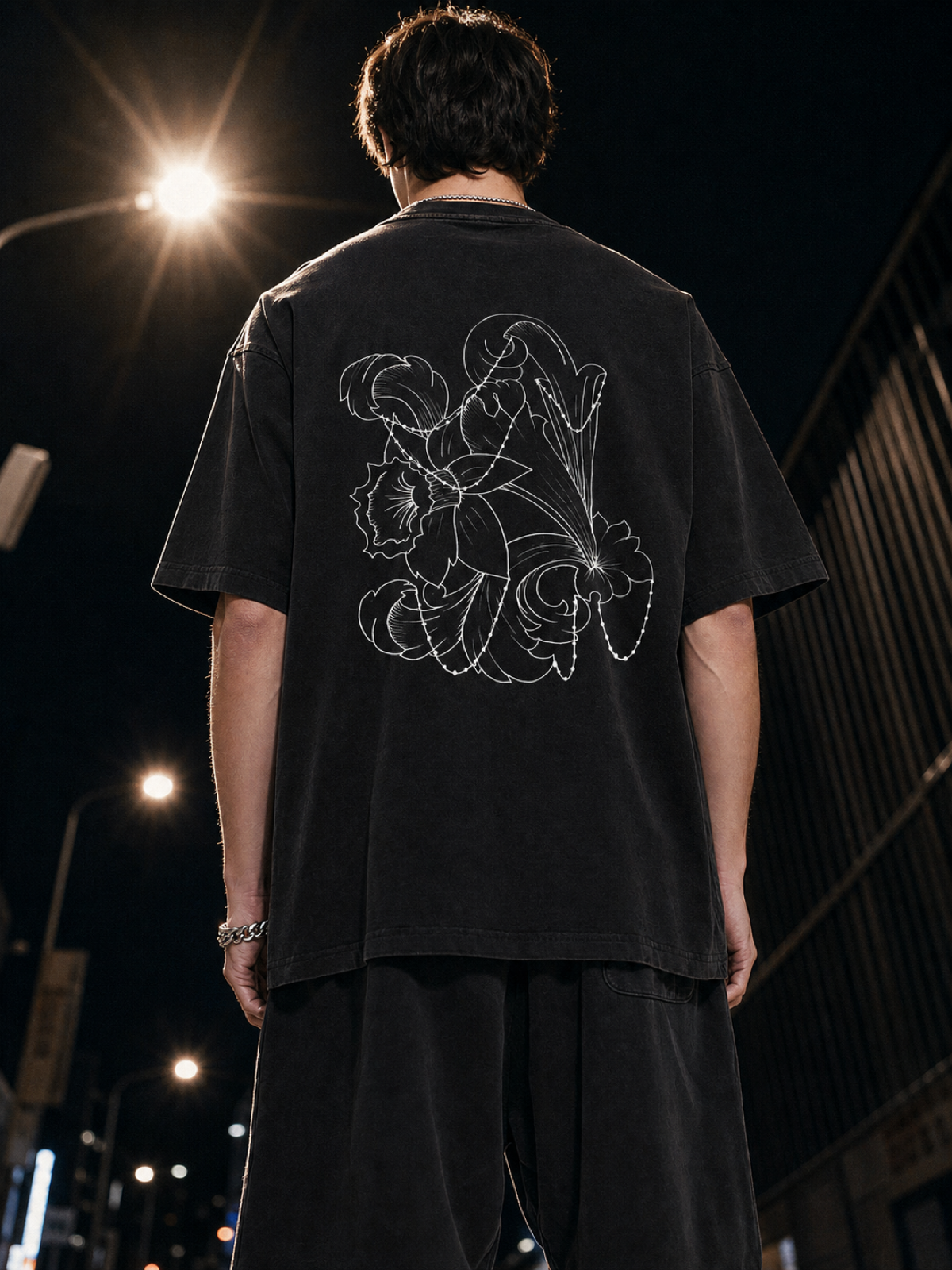 Person wearing a black T-shirt with a white floral and abstract design on the back, standing outdoors at night with streetlights in the background.