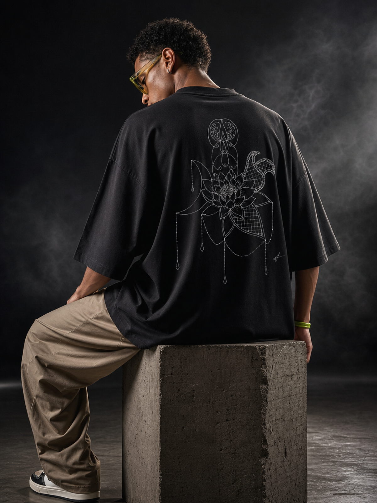 A person with curly hair and glasses sitting on a concrete block, wearing a black oversized T-shirt with a large white embroidered floral and geometric design on the back, beige pants, and black and white sneakers, against a dark background.