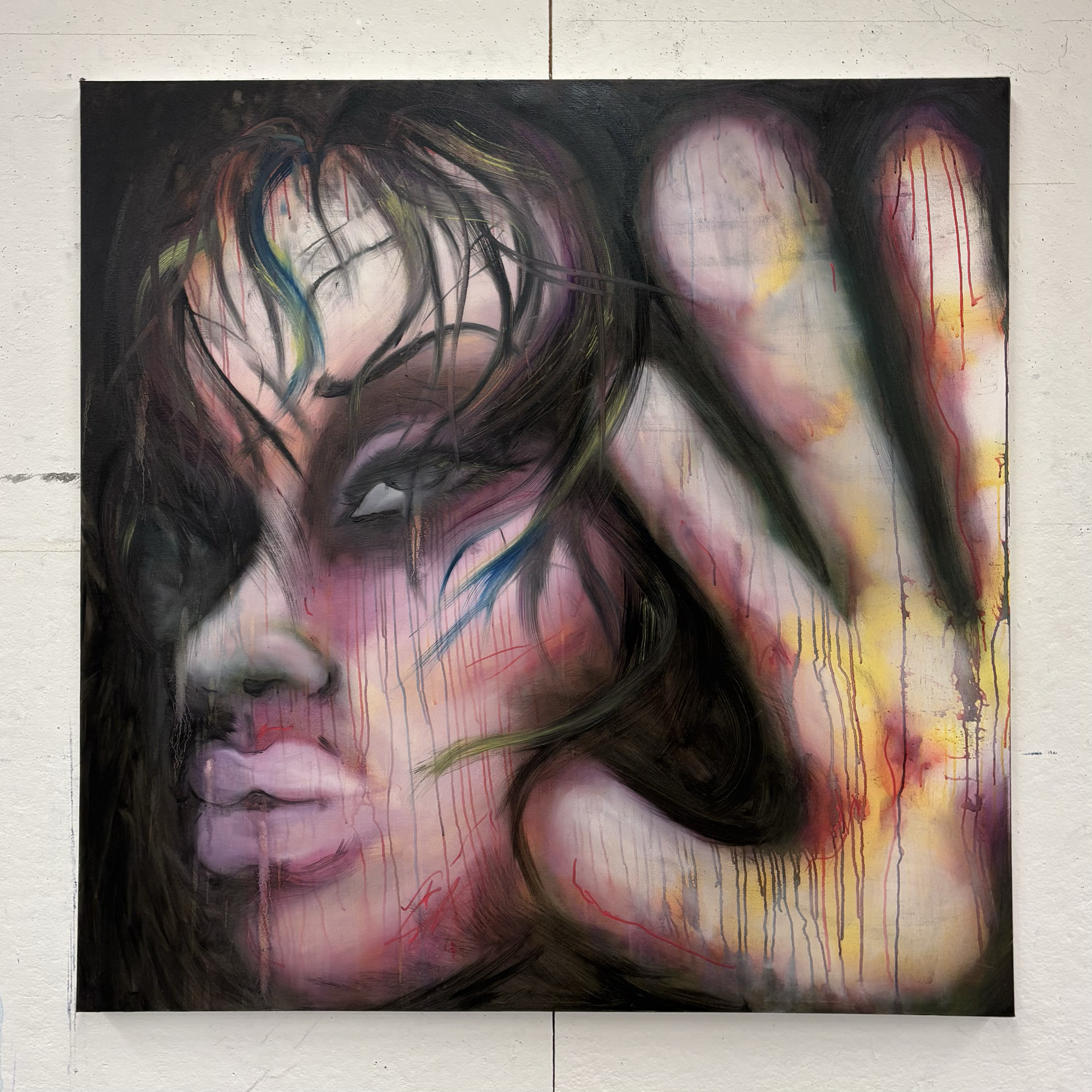 Abstract painting of a woman's face, with dark, flowing hair, in a mix of dark and vibrant colors, with drips of paint, on a white textured wall.