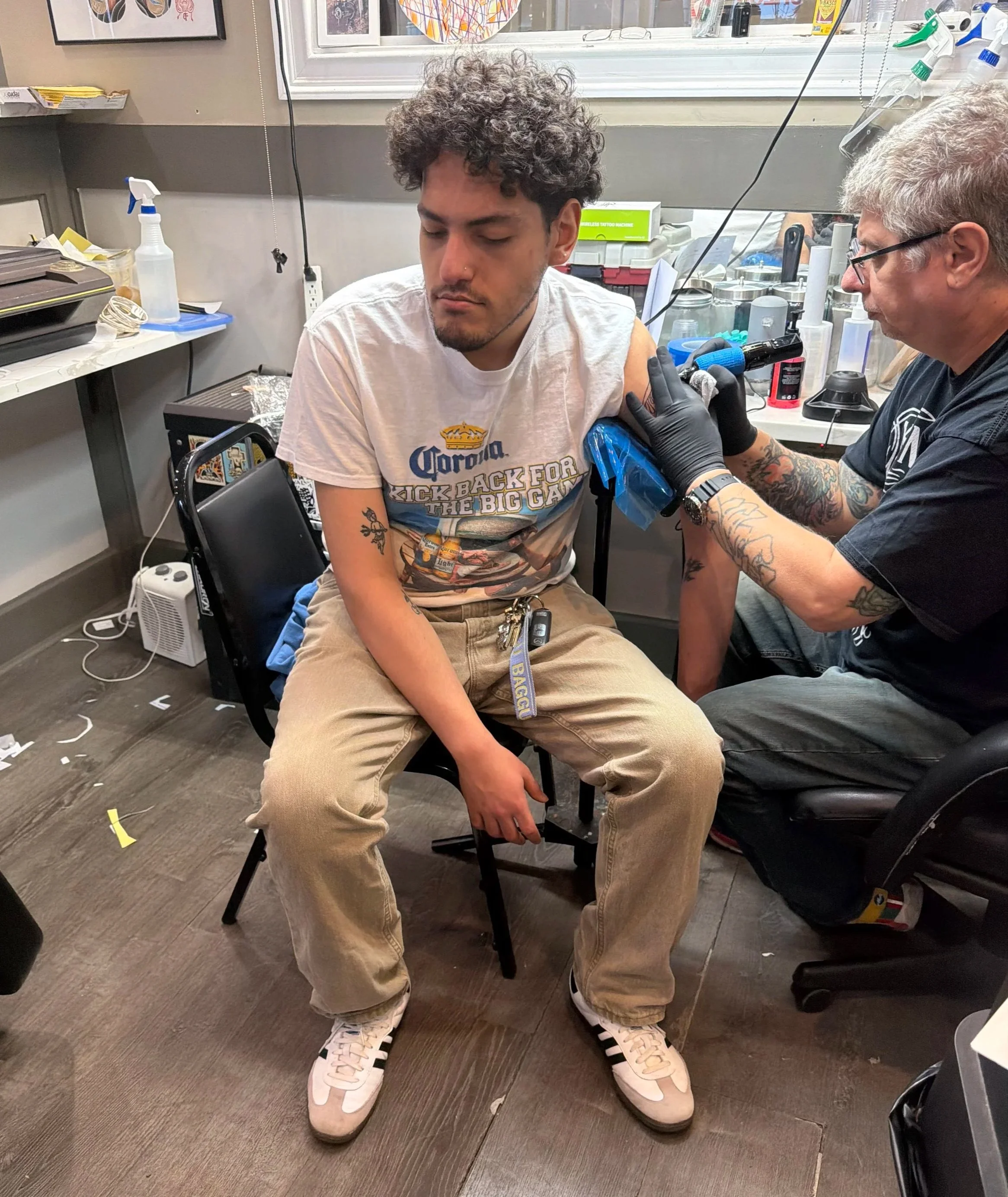 A young man with curly hair and tattoos on his arm getting a tattoo on his upper arm by a tattoo artist in a tattoo studio. The young man has a focused expression, and the artist is wearing gloves and holding a tattoo machine. The studio has various supplies and equipment on the counter, with some colorful artwork on the wall.