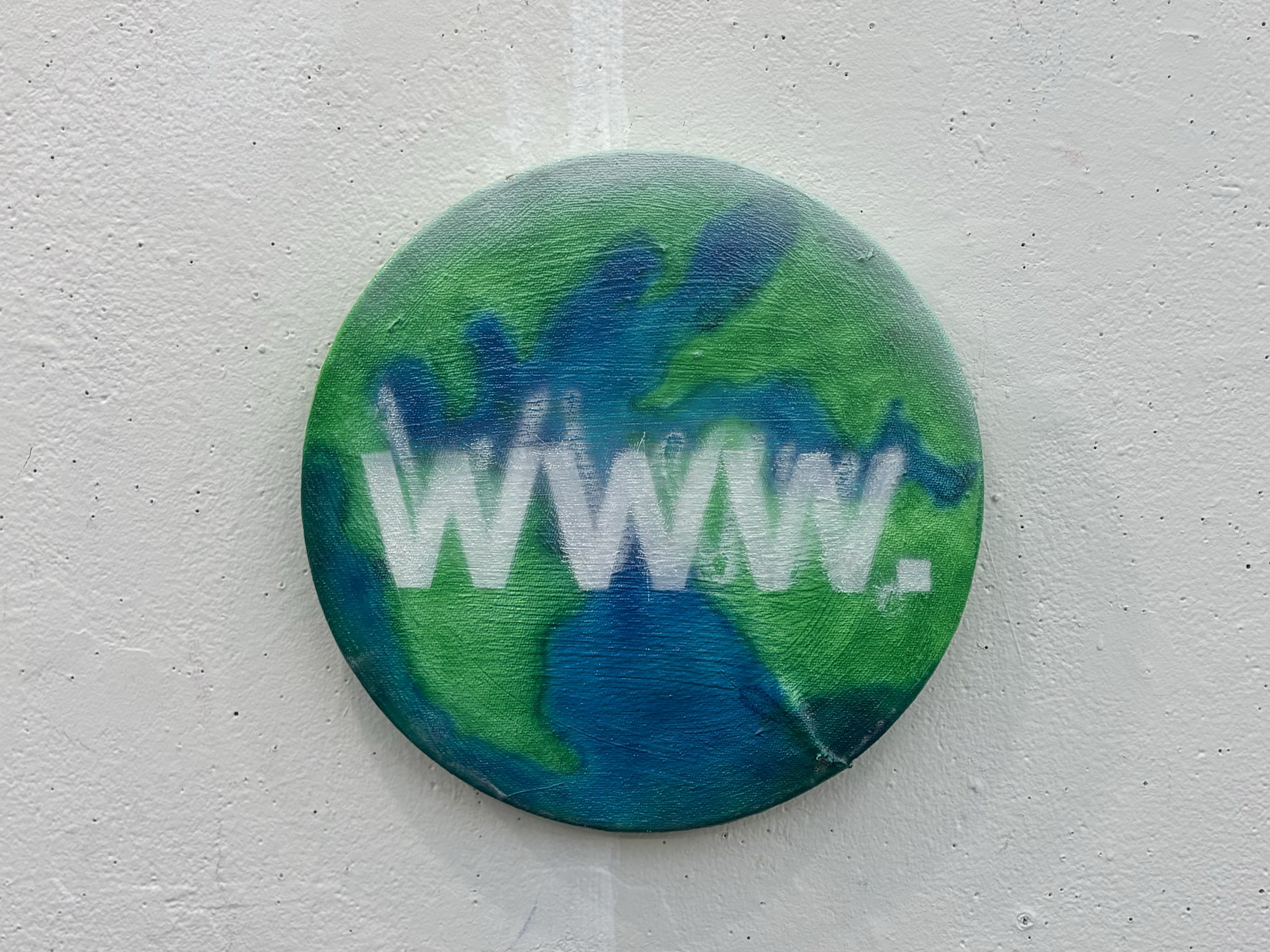 A painted circular object with blue and green colors and white graffiti on top, placed on a textured white wall.