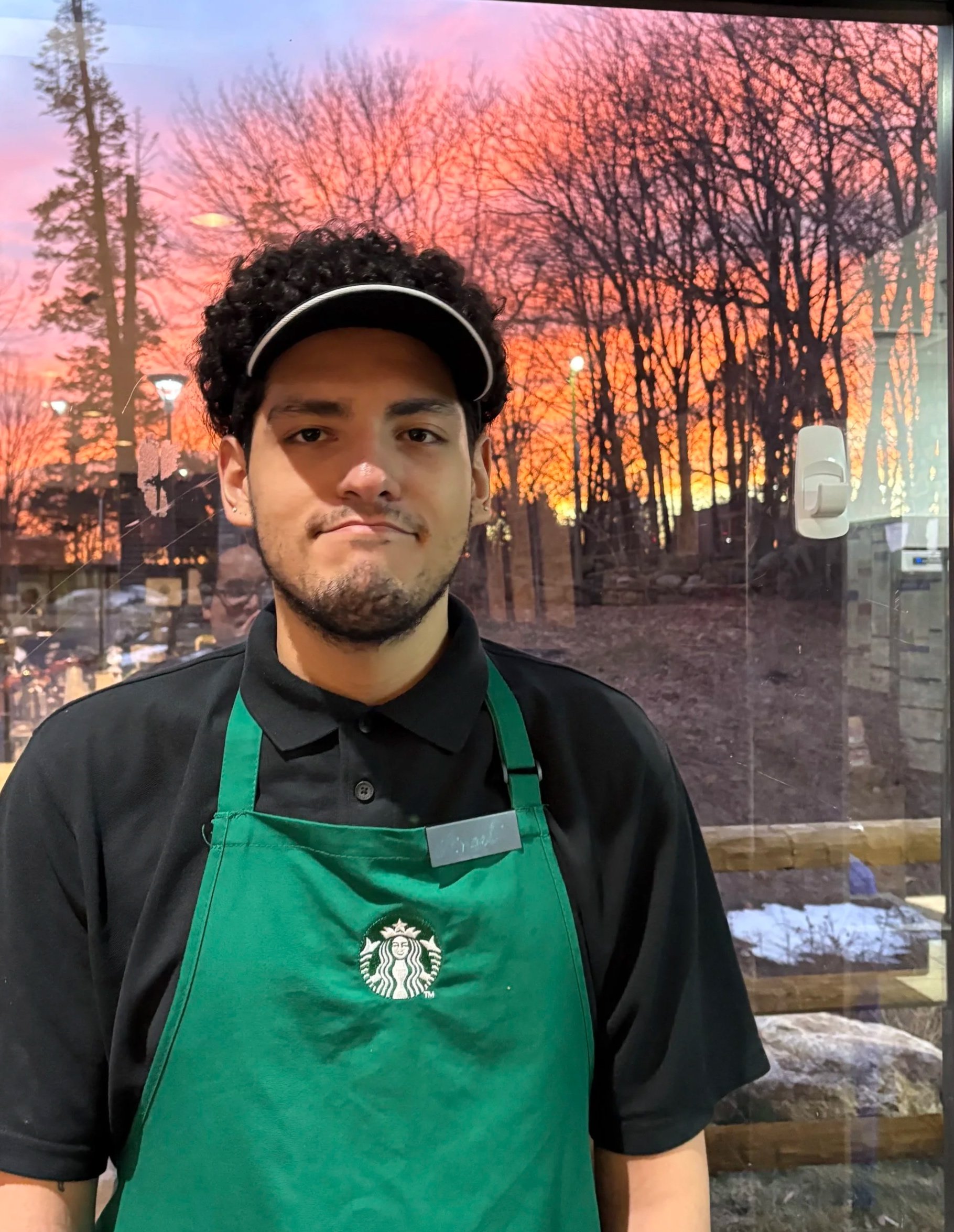 A person with dark curly hair wearing a black visor and black shirt, standing indoors with a view of trees and a sunset sky outside the window behind them. The person is wearing a green Starbucks apron.